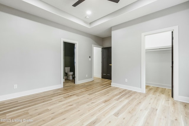 unfurnished bedroom with ensuite bathroom, a spacious closet, light hardwood / wood-style flooring, a tray ceiling, and a closet