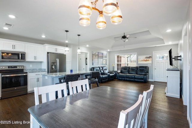 dining area with dark wood-type flooring, ceiling fan with notable chandelier, and a tray ceiling