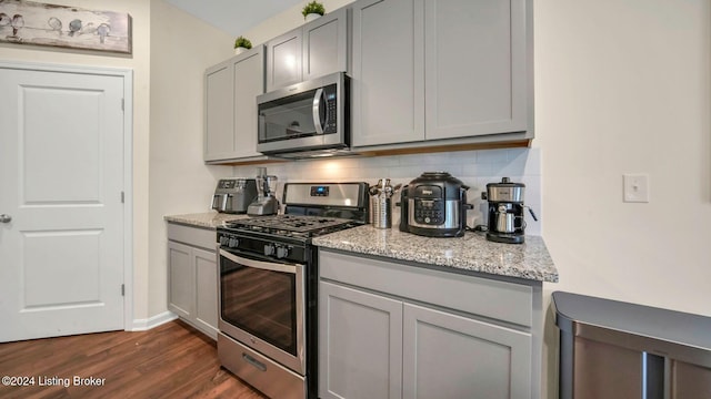 kitchen with appliances with stainless steel finishes, gray cabinetry, light stone counters, dark hardwood / wood-style flooring, and decorative backsplash