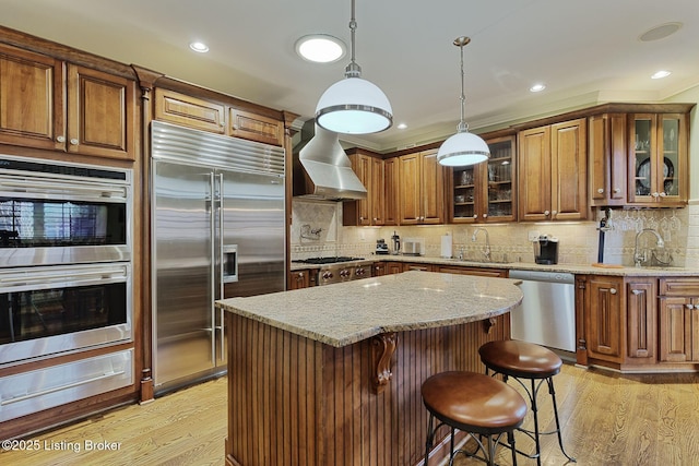 kitchen with stainless steel appliances, a sink, light wood-type flooring, a warming drawer, and wall chimney exhaust hood