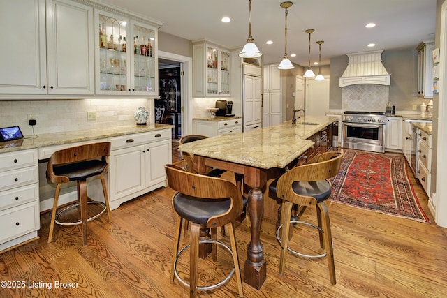 kitchen with pendant lighting, an island with sink, custom exhaust hood, high end stainless steel range, and light stone countertops