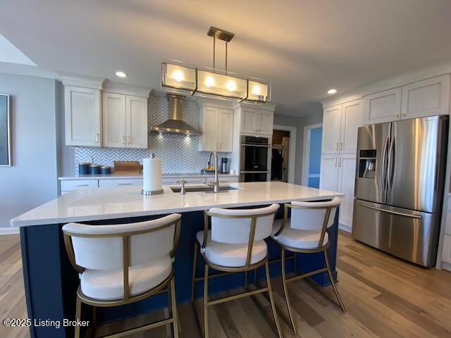 kitchen with appliances with stainless steel finishes, white cabinetry, sink, hanging light fixtures, and wall chimney range hood