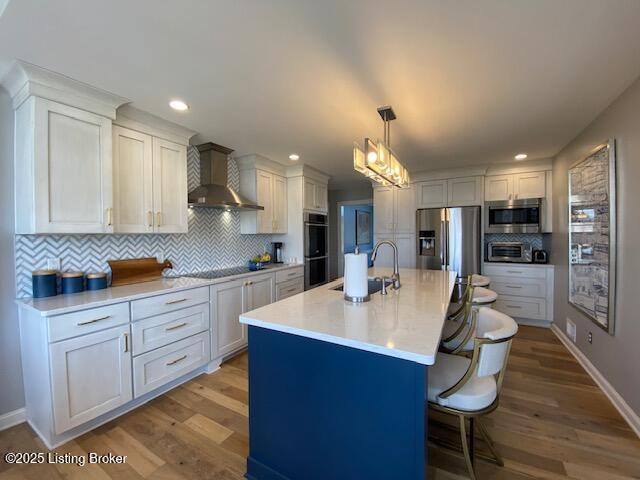 kitchen featuring wall chimney exhaust hood, sink, decorative light fixtures, appliances with stainless steel finishes, and a kitchen island with sink