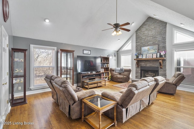 living area with baseboards, ceiling fan, wood finished floors, a stone fireplace, and high vaulted ceiling