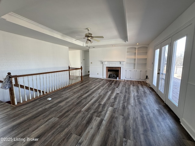 unfurnished living room featuring crown molding, dark wood-type flooring, built in features, ceiling fan, and a tray ceiling