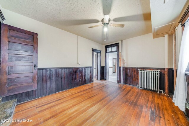 spare room with a wainscoted wall, a textured ceiling, wood-type flooring, radiator, and ceiling fan