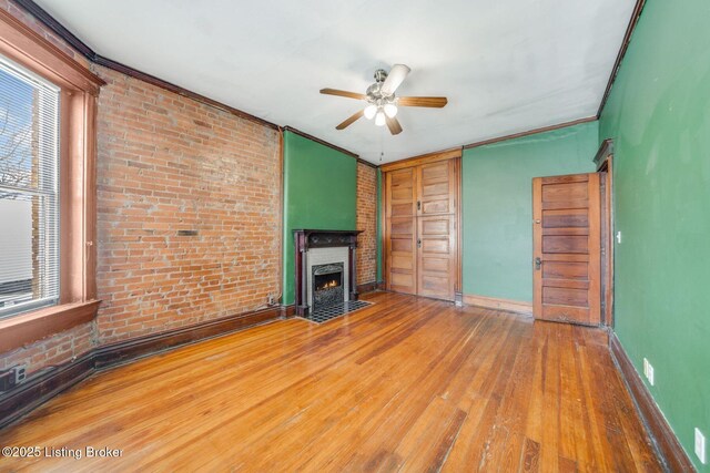 unfurnished living room with a fireplace with flush hearth, wood finished floors, brick wall, crown molding, and baseboards