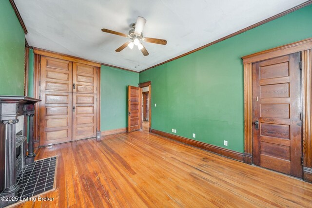 unfurnished bedroom with a tiled fireplace, wood finished floors, crown molding, baseboards, and ceiling fan