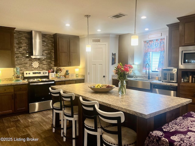 kitchen with pendant lighting, appliances with stainless steel finishes, wall chimney exhaust hood, and a kitchen island