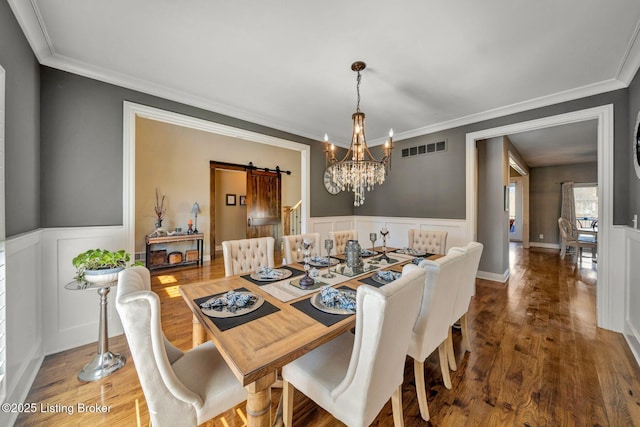 dining space featuring wood finished floors, a wainscoted wall, visible vents, ornamental molding, and a barn door