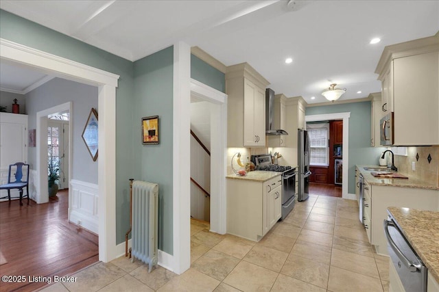 kitchen featuring radiator, wall chimney range hood, sink, appliances with stainless steel finishes, and decorative backsplash