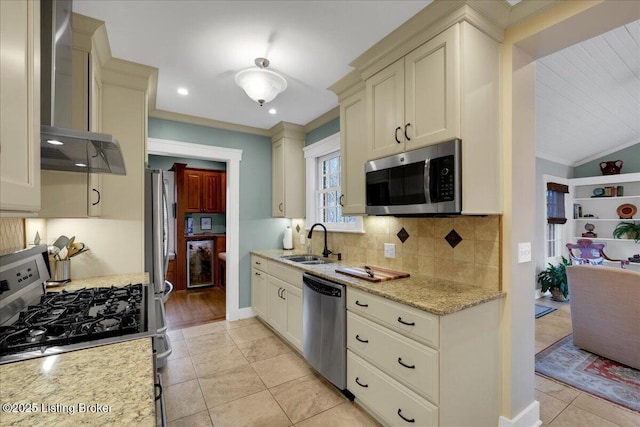 kitchen with sink, appliances with stainless steel finishes, light stone countertops, cream cabinets, and wall chimney range hood