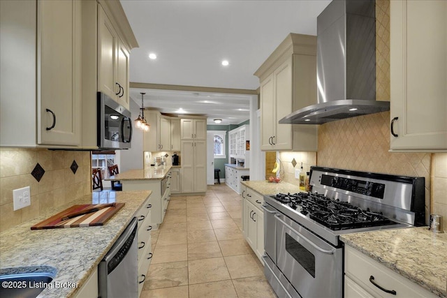 kitchen with appliances with stainless steel finishes, cream cabinets, decorative light fixtures, light stone countertops, and wall chimney range hood