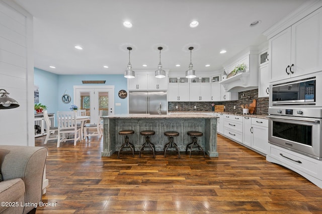 kitchen featuring dark wood-style flooring, glass insert cabinets, white cabinetry, built in appliances, and a kitchen bar