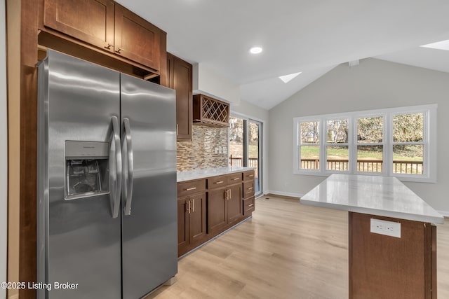 kitchen with backsplash, vaulted ceiling with skylight, stainless steel refrigerator with ice dispenser, light hardwood / wood-style floors, and a kitchen island