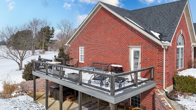 snow covered rear of property featuring a shingled roof, brick siding, and a deck