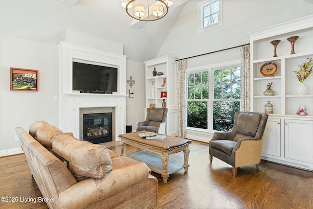 living area with baseboards, dark wood finished floors, a glass covered fireplace, an inviting chandelier, and high vaulted ceiling