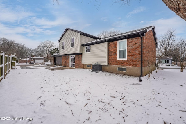 snow covered back of property with a garage, central AC, brick siding, and fence