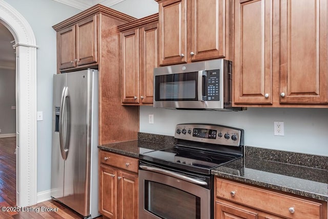 kitchen featuring arched walkways, stainless steel appliances, brown cabinetry, wood finished floors, and dark stone counters