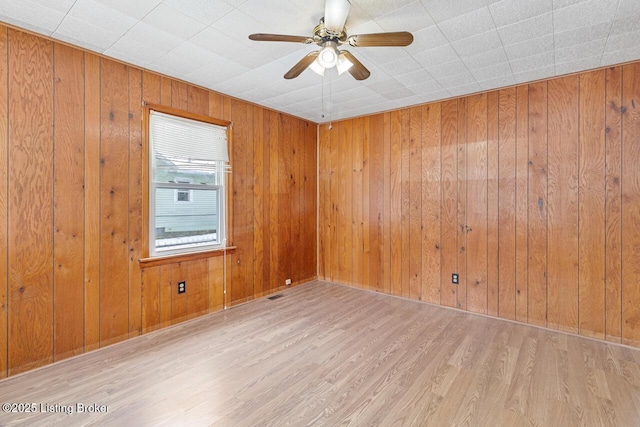 empty room with light wood-style flooring, wooden walls, ceiling fan, and visible vents