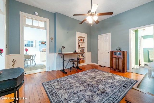 home office featuring wainscoting, wood finished floors, a ceiling fan, and built in features