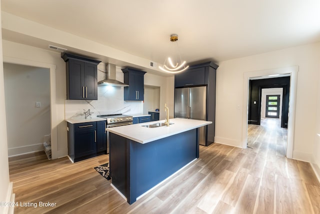 kitchen with a kitchen island with sink, stainless steel appliances, a sink, light countertops, and wall chimney range hood