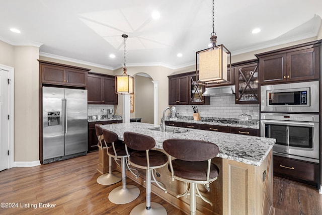 kitchen with arched walkways, dark wood-style flooring, a center island with sink, appliances with stainless steel finishes, and a kitchen bar