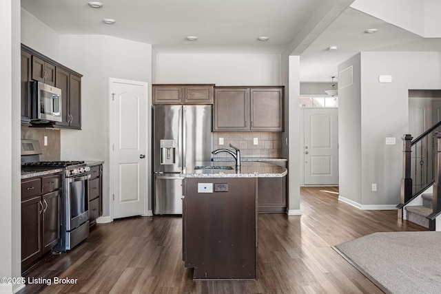 kitchen featuring an island with sink, appliances with stainless steel finishes, light stone counters, dark wood-type flooring, and a sink