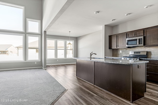 kitchen with dark brown cabinetry, dark wood-type flooring, a kitchen island with sink, stainless steel appliances, and backsplash