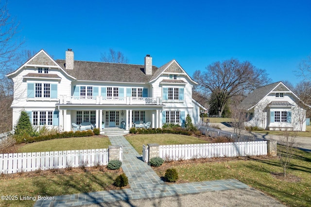 shingle-style home featuring covered porch, a fenced front yard, a chimney, and a balcony