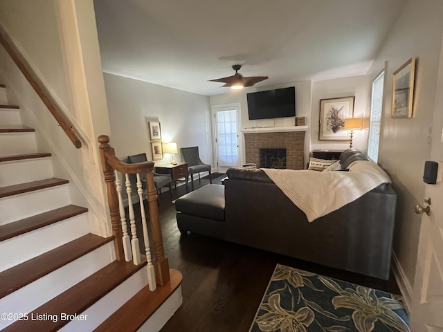 living room featuring a brick fireplace, stairs, a ceiling fan, and dark wood-style flooring