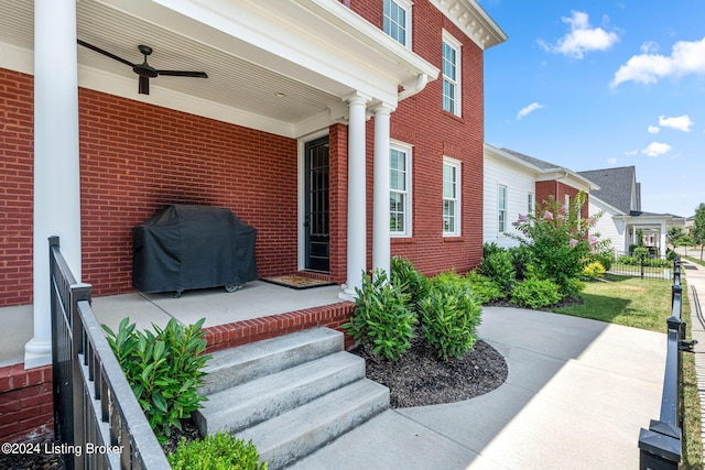 view of exterior entry with a ceiling fan, covered porch, brick siding, and fence