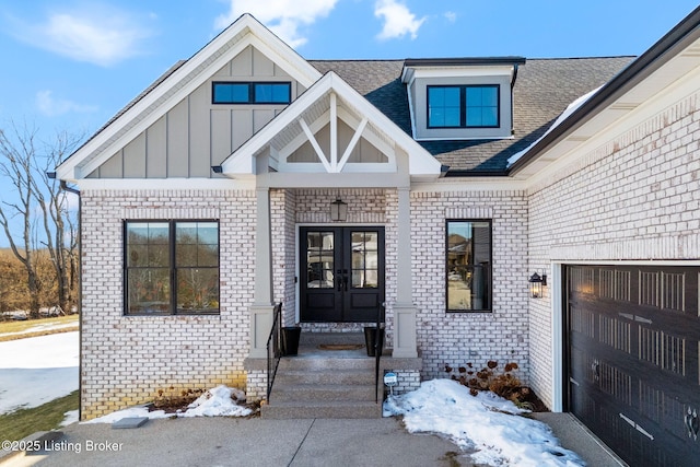 snow covered property entrance with a garage, brick siding, french doors, roof with shingles, and board and batten siding