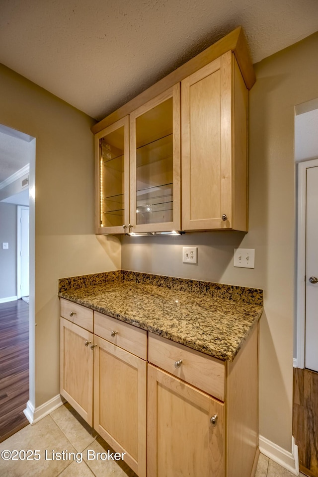 kitchen featuring baseboards, glass insert cabinets, stone counters, light brown cabinets, and light tile patterned flooring