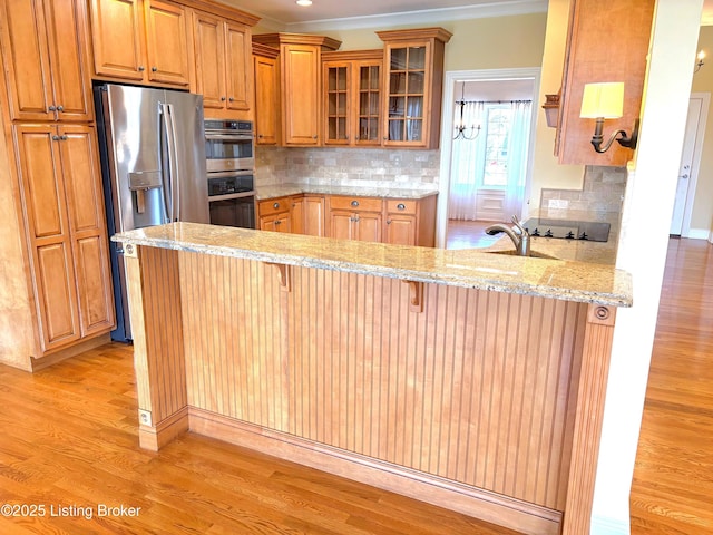 kitchen with light wood-type flooring, appliances with stainless steel finishes, a peninsula, glass insert cabinets, and light stone countertops