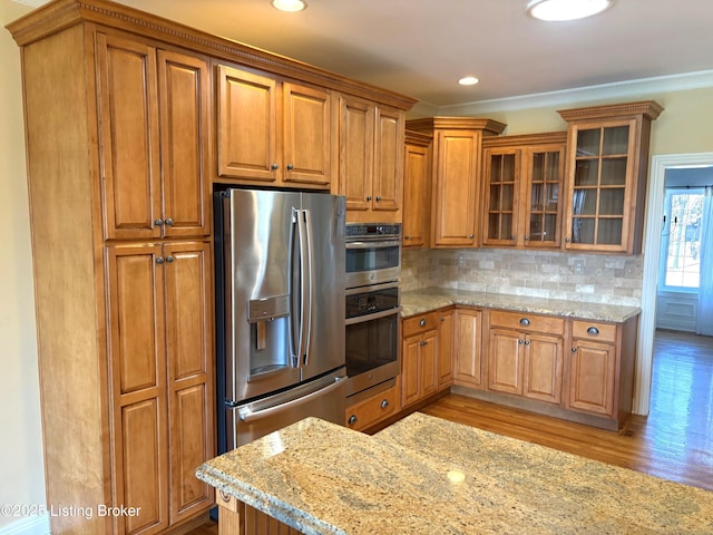 kitchen with stainless steel appliances, brown cabinetry, light wood finished floors, glass insert cabinets, and light stone countertops