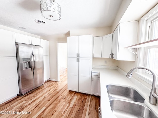 kitchen with stainless steel refrigerator with ice dispenser, light wood finished floors, light countertops, white cabinets, and a sink