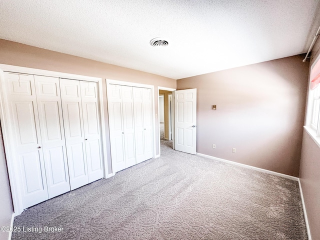 unfurnished bedroom with a textured ceiling, carpet floors, visible vents, baseboards, and two closets