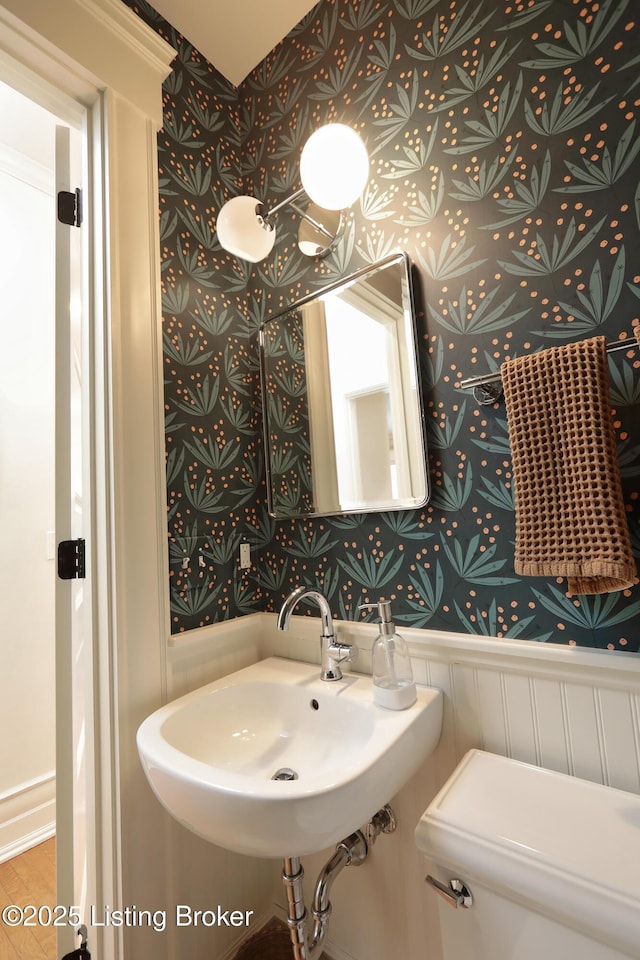 bathroom with a wainscoted wall, a sink, toilet, and wallpapered walls