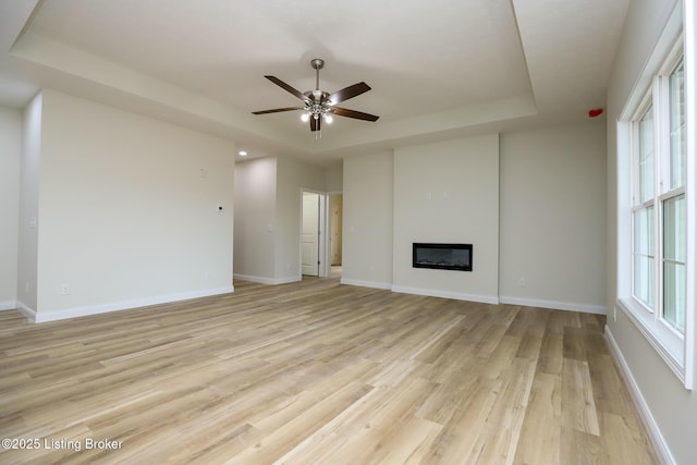 unfurnished living room with a glass covered fireplace, a raised ceiling, light wood-style floors, and baseboards