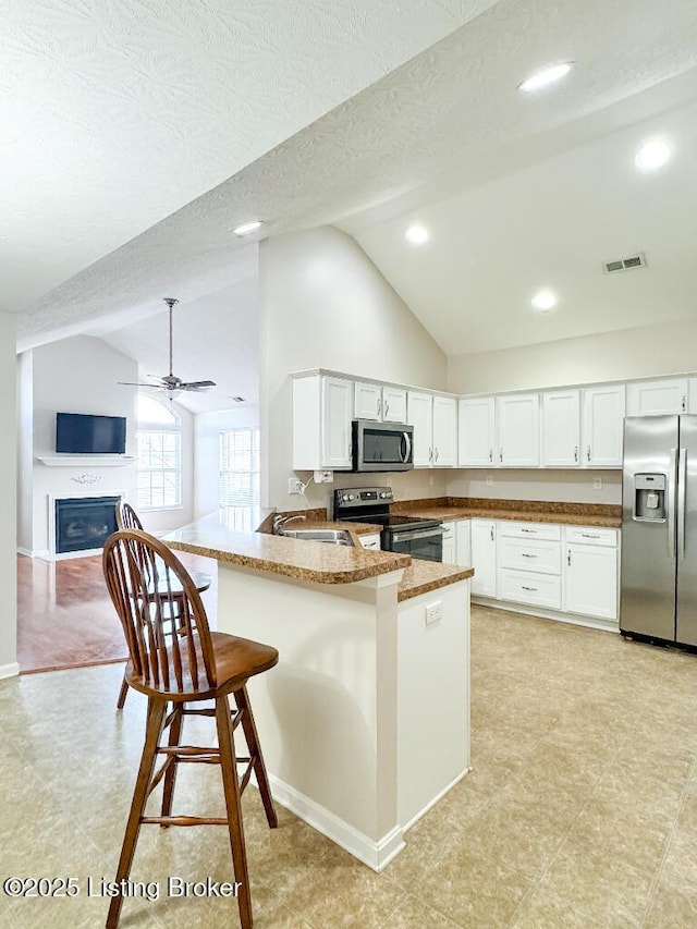 kitchen with a peninsula, a fireplace, white cabinetry, appliances with stainless steel finishes, and a kitchen bar