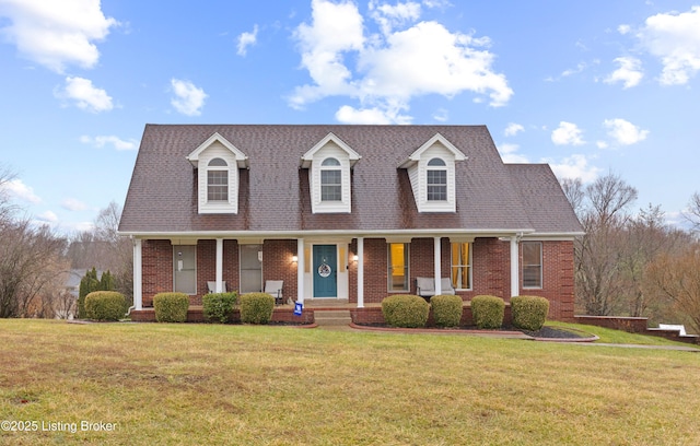 new england style home featuring a shingled roof, a front yard, covered porch, and brick siding