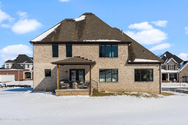 snow covered rear of property featuring french doors, brick siding, and roof with shingles