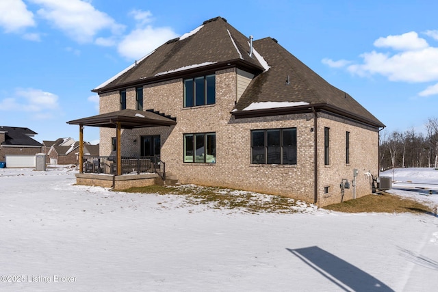 snow covered house featuring a porch, brick siding, a shingled roof, and central air condition unit