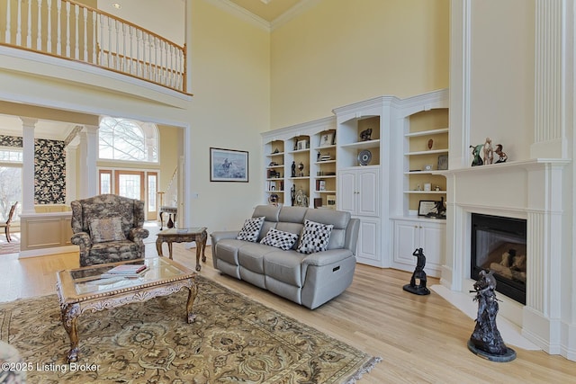 living room with a towering ceiling, ornamental molding, light wood finished floors, a glass covered fireplace, and ornate columns