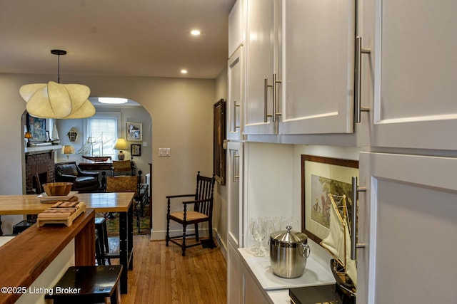 kitchen featuring arched walkways, light wood-style flooring, a brick fireplace, white cabinetry, and pendant lighting