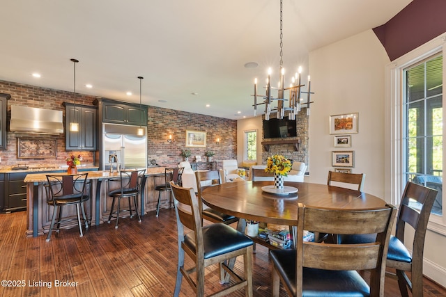 dining room with a chandelier, a stone fireplace, recessed lighting, brick wall, and dark wood finished floors