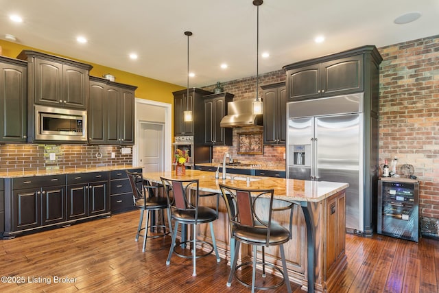 kitchen with beverage cooler, an island with sink, wall chimney exhaust hood, built in appliances, and hanging light fixtures