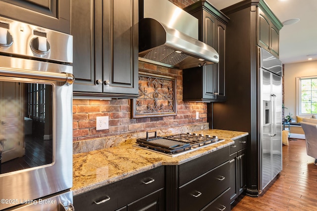 kitchen with dark brown cabinetry, light wood finished floors, light stone countertops, stainless steel appliances, and wall chimney range hood