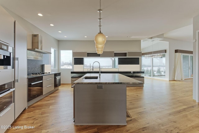 kitchen featuring a kitchen island with sink, oven, a sink, light wood-type flooring, and wall chimney exhaust hood